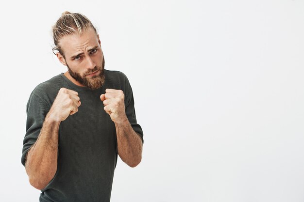 Portrait of attractive young man with trendy hairstyle and beard holding hands in front of him in boxing position going to fight.