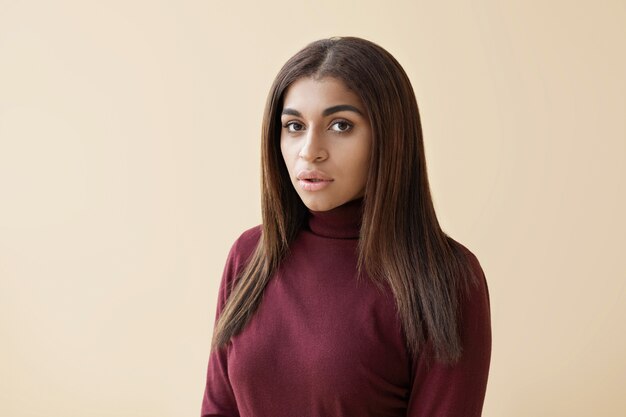 Portrait of attractive young mixed race lady with long hair dressed in maroon turtleneck sweater standing
