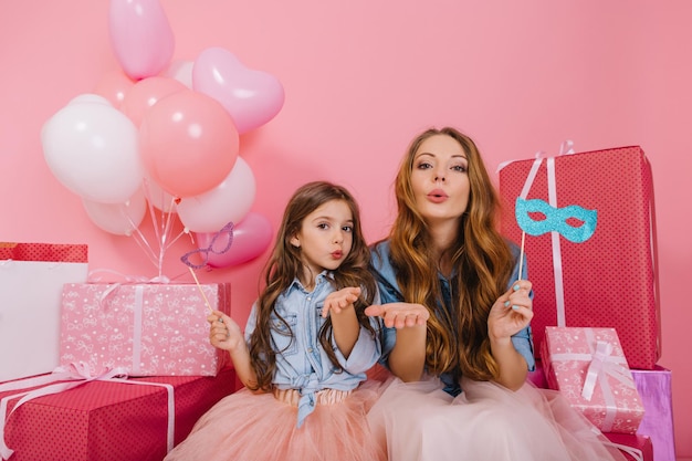 Portrait of attractive young mother and cute daughter sending air kisses after birthday party. Two adorable sisters with lovely faces posing before opening gifts, sitting with colorful balloons