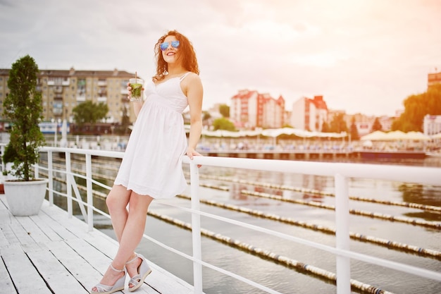 Ritratto di una giovane donna attraente in posa con il suo cocktail su una riva del lago che indossa occhiali da sole