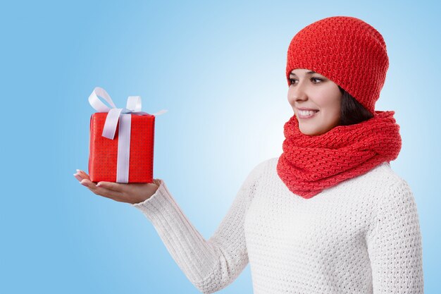 Portrait of an attractive young woman standing sideways on blue wearing warm winter clothes holding a present in her hand