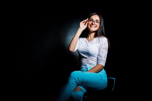 Portrait of an attractive young woman in white top and blue pants sitting posing with her glasses in the dark