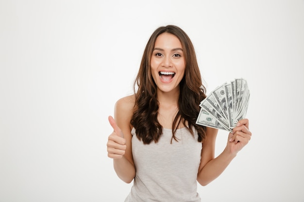 Portrait of attractive young woman with long hair holding lots of money cash, smiling on camera showing thumb up over white wall