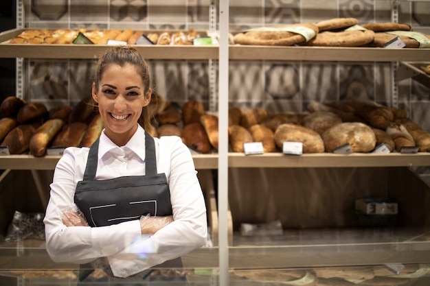 Portrait of bakery seller with arms crossed standing in front of shelf full of bred bagels and pastry