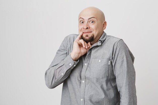 Portrait of bald bearded man with funny expression holding his pinky near mouth on white