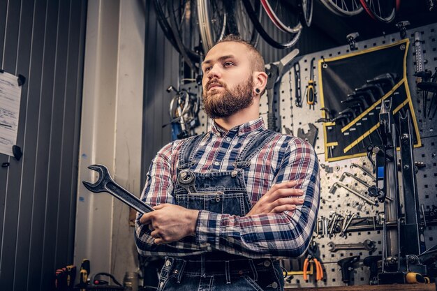 Portrait of bearded bicycle mechanic with crossed arms holds cup key over tool stand background in a workshop.