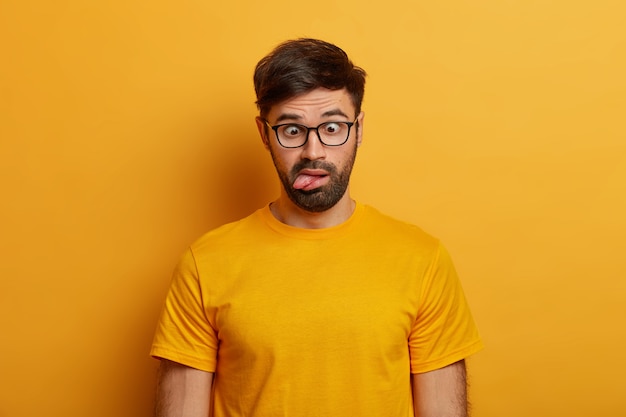 Portrait of bearded guy shows grimace, crosses eyes and sticks out tongue, plays around, going crazy, wears spectacles, everyday t shirt, poses against yellow wall. Human face expressions