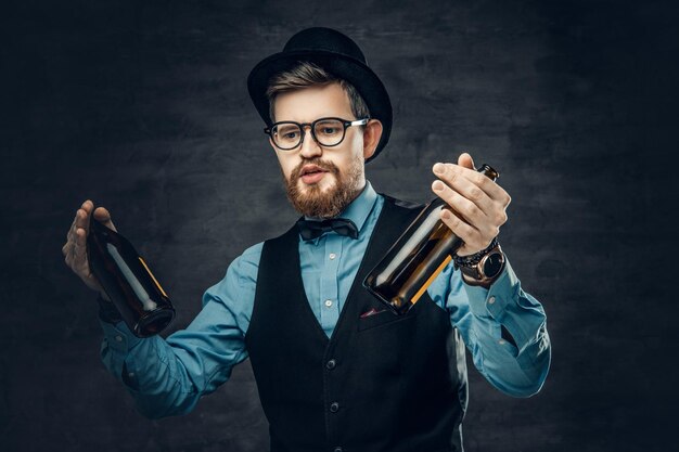 Portrait of bearded hipster male dressed in a blue shirt, elegant waistcoat and top hat holds two craft beer bottles with a think of a party.