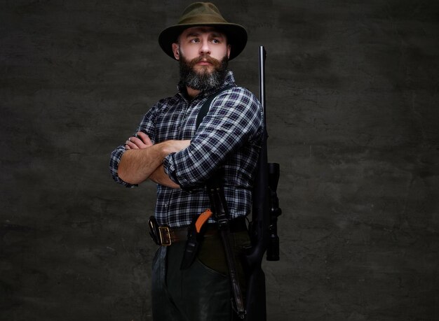 Portrait of a bearded hunter in a fleece shirt and hat standing with a rifle behind his back. Isolated on a dark background.