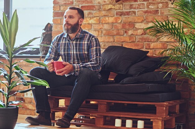 Portrait of bearded male dressed in a blue fleece shirt, drinks hot coffee near the window in the rest room with loft interior.