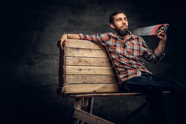 Free photo portrait of bearded male dressed in a jeans and a plaid shirt holds handsaw on a shoulder.