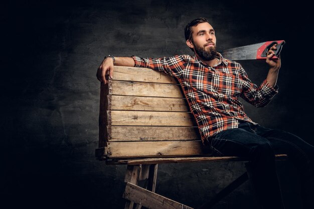 Portrait of bearded male dressed in a jeans and a plaid shirt holds handsaw on a shoulder.