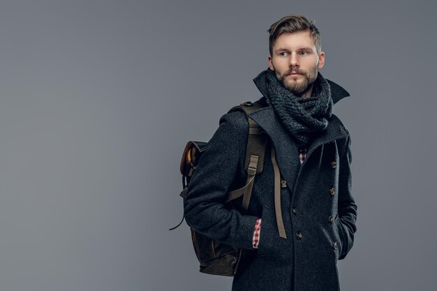 Portrait of bearded male dressed in a warm jacket and a scarf  hold a backpack isolated on grey background.