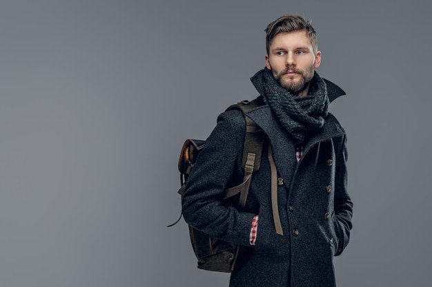 Портрет бородатого мужчины, одетого в теплую куртку и шарф, держит рюкзак на сером фоне.