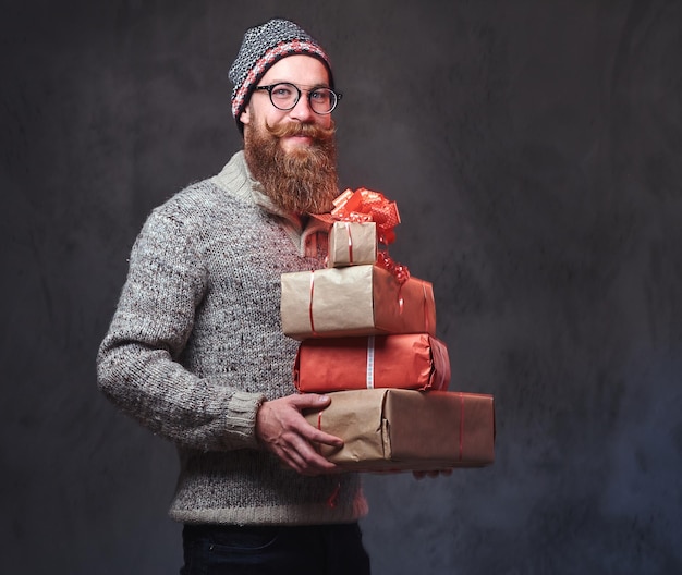 따뜻한 모직 스웨터와 모자를 쓴 안경을 쓴 수염 난 남성의 초상화는 회색 배경 위에 크리스마스 선물을 들고 있습니다.