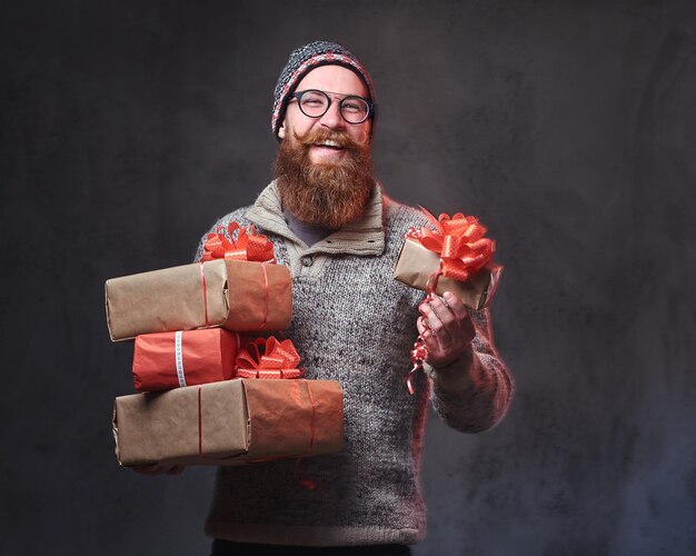 Portrait of bearded male in eyeglasses dressed in a warm wool sweater and a hat holds Christmas gifts over grey background.