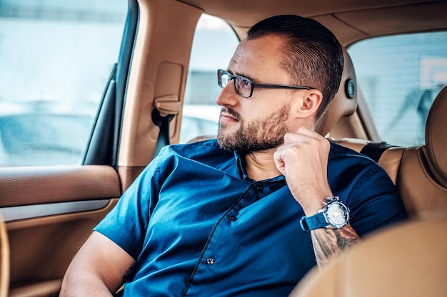 Portrait of bearded male in eyeglasses with tattoo on his arms, sits sits in a car.