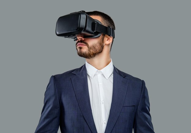 Portrait of bearded male in a suit with virtual reality glasses on his head isolated on grey background.