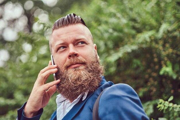 Portrait of a bearded male with a haircut dressed in a shirt and jacket with a backpack, talking by phone in a park.