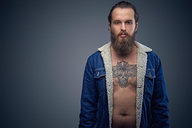 Foto gratuita ritratto di uomo barbuto con un tatuaggio sul petto, vestito con una giacca di jeans su sfondo grigio.