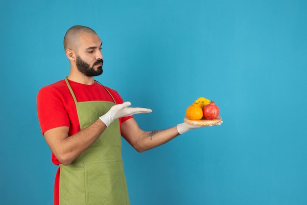 Portrait of a bearded man in apron holding a wooden board of fresh fruits . 