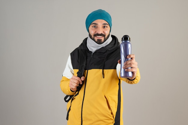 Portrait of bearded man in warm hat standing and holding water bottle against gray wall . 