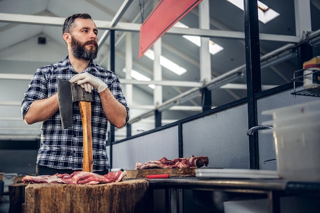 フリースシャツを着たひげを生やした肉の男の肖像画は、斧と新鮮なカット肉を保持しています。