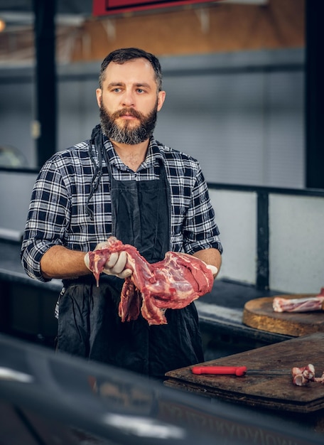 フリースシャツを着たひげを生やした肉の男の肖像画は、市場で新鮮なカット肉を保持しています。