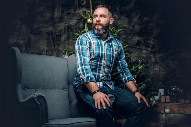 Портрет бородатого мужчины средних лет, одетого в клетчатую фланелевую рубашку и джинсы, сидит на стуле на рождественском фоне.