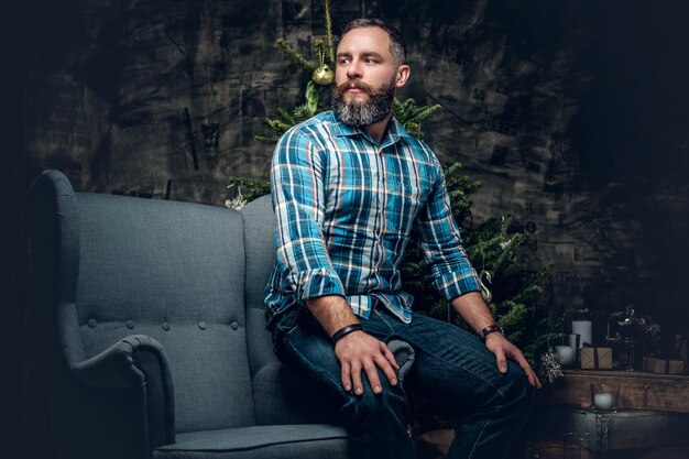 Portrait of bearded middle age male dressed in plaid flannel shirt and jeans sits on a chair over Christmas decorated background.