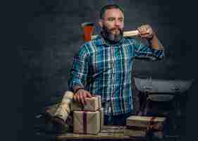 Free photo portrait of bearded middle age male holds an axe and standing near a table with christmas gift boxes and firewoods over grey background.