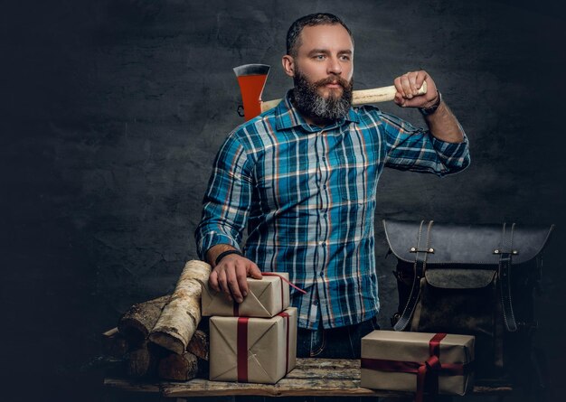 Portrait of bearded middle age male holds an axe and standing near a table with Christmas gift boxes and firewoods over grey background.