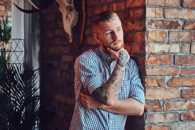 Portrait of a bearded modern male with tattoos on his arms posing near the window in a room with loft interior.