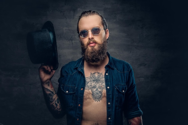 Portrait of bearded tattooed hipster male wearing a top hat over grey vignette background.