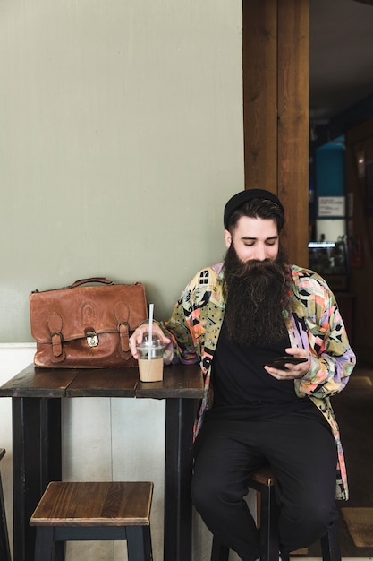 Foto gratuita ritratto di un giovane uomo barbuto seduto nella caffetteria con il cellulare