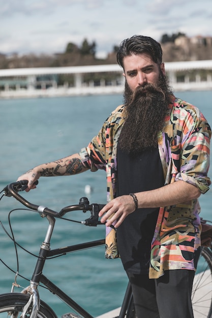 Free photo portrait of bearded young man standing with his bicycle looking at camera