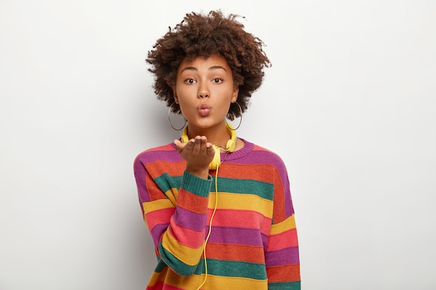 Portrait of beautiful affectionate curly teenage girl keeps palms stretched forward, lips rounded, sends air kiss, wears casual colorful jumper, uses headphones for listening favourite melodies