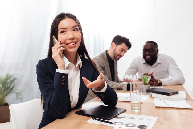 Portrait of beautiful asian businesswoman sitting in office and joyfully looking aside while talking on her cellphone with multinational business partners on background