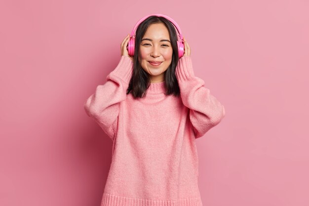 Portrait of beautiful Asian female meloman wears wireless electronic stereo headpones listens favorite audio track or popular song recreats with good music enjoys tranquil melody wears pink sweater