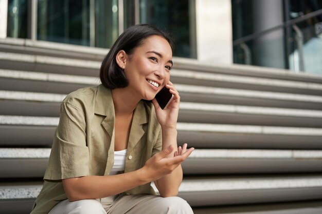 Портрет красивой азиатской девушки разговаривает по мобильному телефону сидит на уличной лестнице Женщина со смартфоном улыбается, звонит