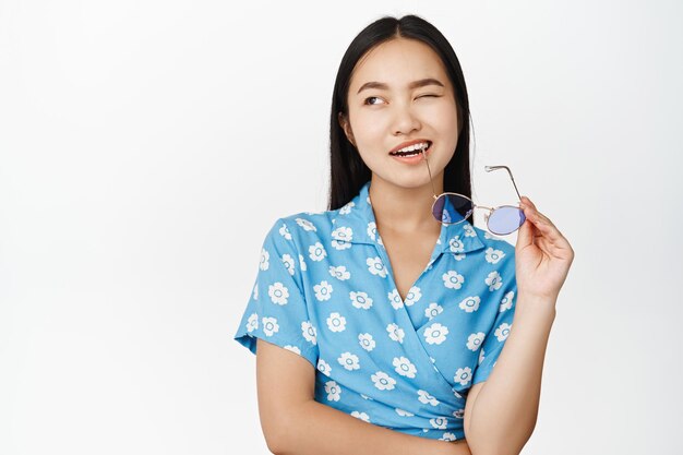 Portrait of beautiful asian modern girl biting temple of sunglasses winking and smiling with confidence standing over white background