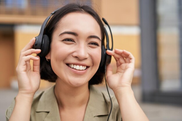 Free photo portrait of beautiful asian woman in headphones listening music on street of city centre smiling