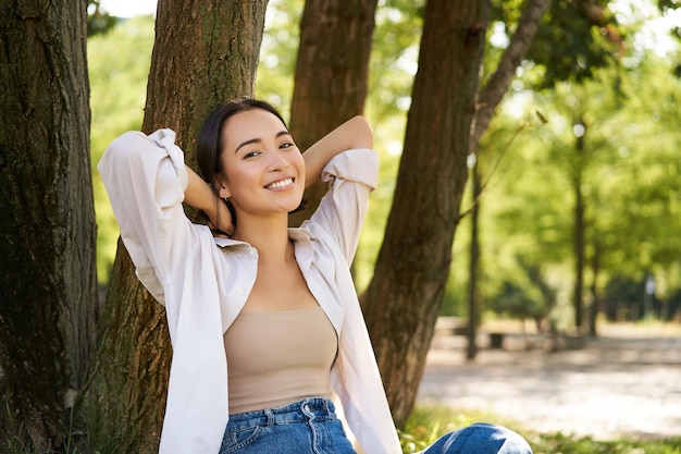 Foto gratuita ritratto di bella donna asiatica che riposa vicino all'albero che si rilassa nel parco sorridendo e sembrando felice