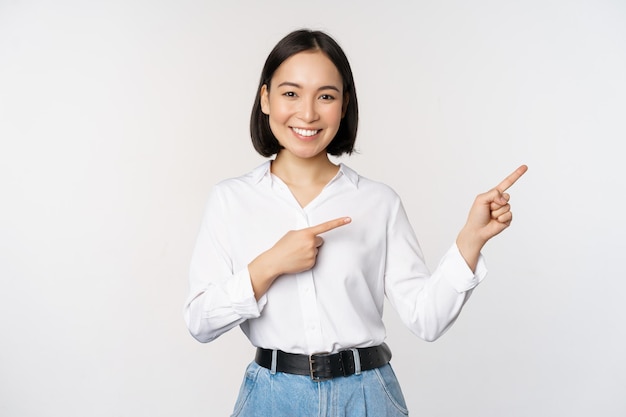 손가락을 오른쪽으로 가리키고 흰색 배경 위에 서 있는 정보 배너 또는 로고 판매 광고를 보여주는 아름다운 아시아 여성 판매원의 초상화