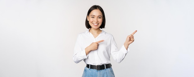 손가락을 오른쪽으로 가리키고 흰색 배경 위에 서 있는 정보 배너 또는 로고 판매 광고를 보여주는 아름다운 아시아 여성 판매원의 초상화