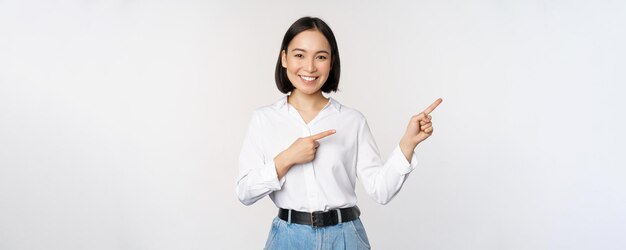 Portrait of beautiful asian woman saleswoman pointing fingers right and showing info banner or logo sale advertisement standing over white background