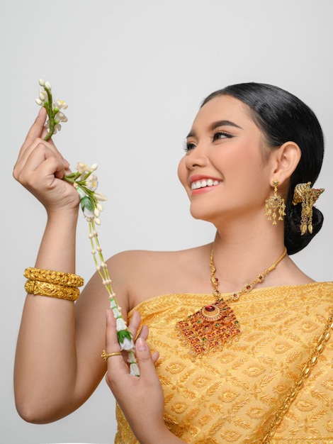 Портрет красивой азиатской женщины в традиционном тайском платье, костюм улыбается и держит гирлянду с изящной позой на белой стене
