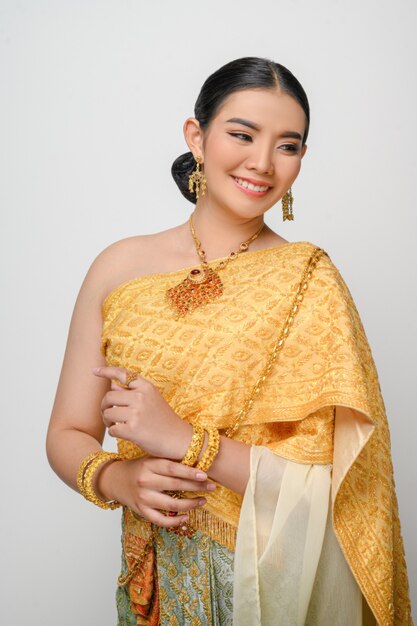 태국 전통 의상을 입은 아름다운 아시아 여성이 흰 벽에 우아하게 미소 짓고 포즈를 취합니다.