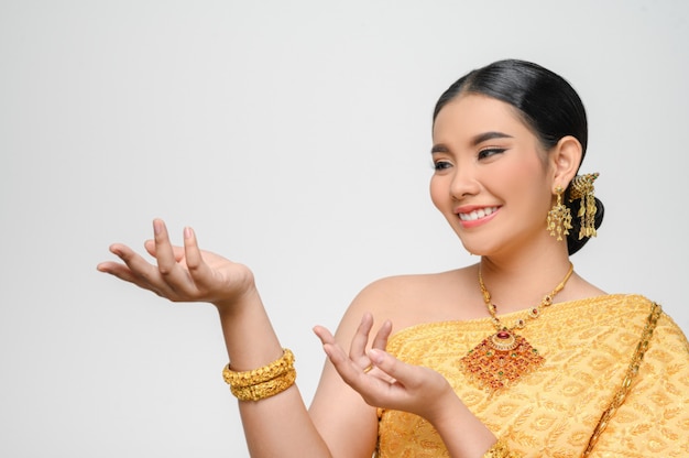 태국 전통 의상을 입은 아름다운 아시아 여성이 하얀 벽에 우아하게 열린 손바닥으로 미소를 짓고 포즈를 취합니다.