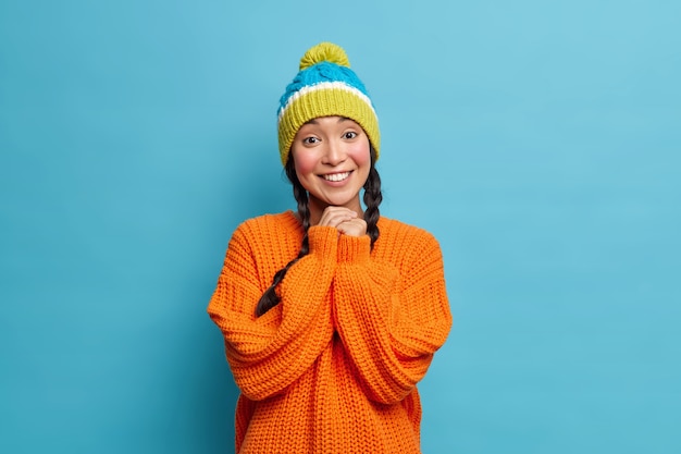 Portrait of beautiful Asian woman with toothy smile keeps hands together being in good mood wears winter knitted hat and orange sweater poses against blue wall
